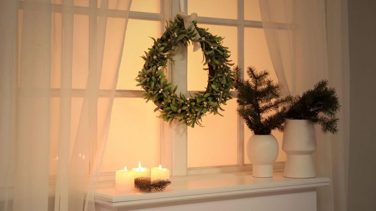 Vases with Christmas branches and candles on fireplace near window in room