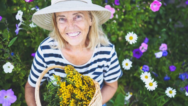 a smiling woman with hat holds basket full of yarrow in a flower garden