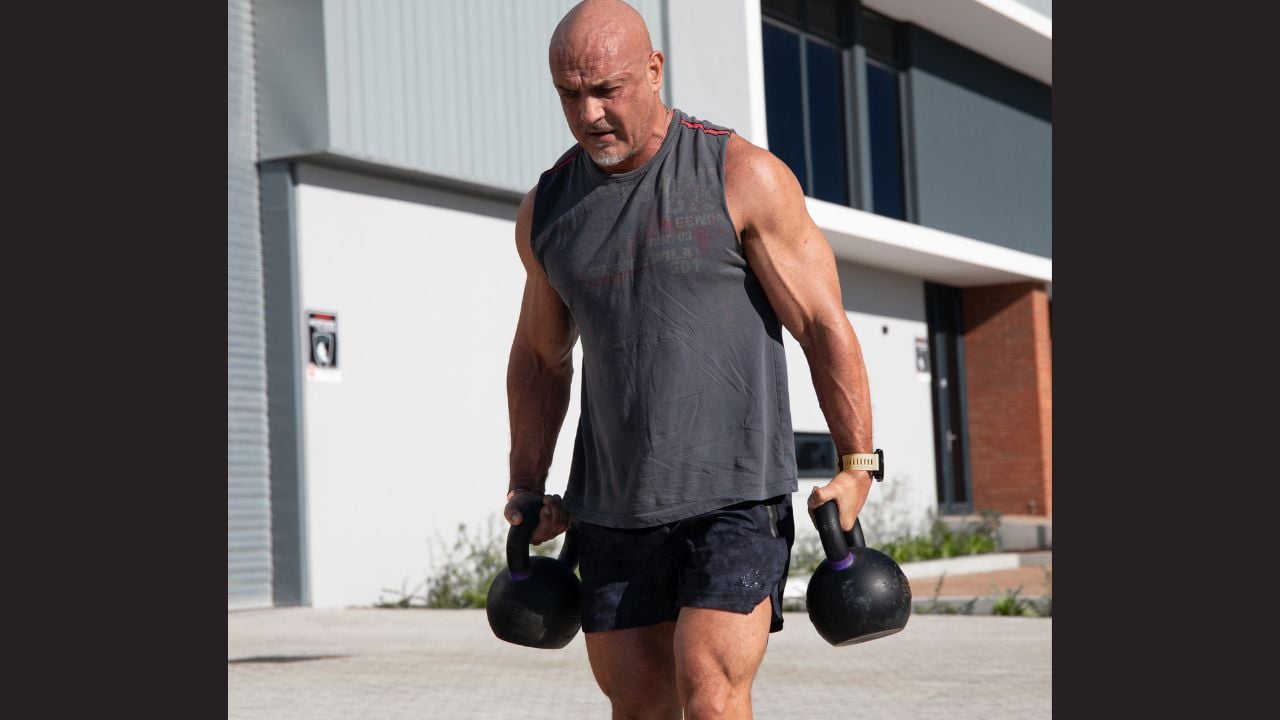 A bald muscled guy in muscle T and rugby shorts walking with heavy kettle bells.