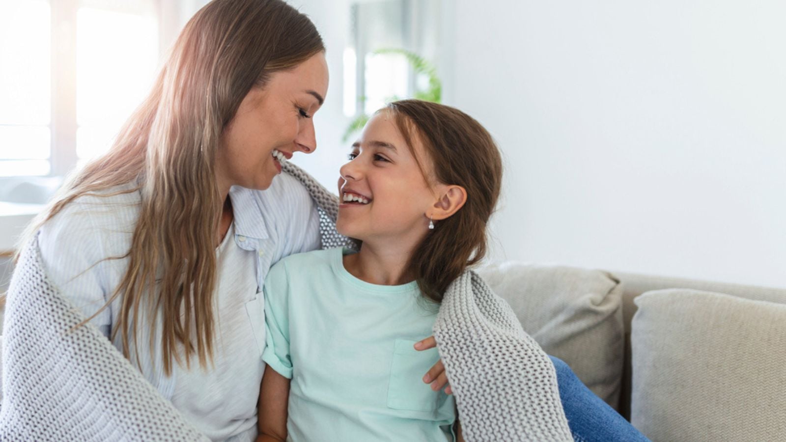 A nice girl and her mother enjoy sunny morning. Good time at home. Child wakes up from sleep. Family playing under blanket on the bed in the bedroom