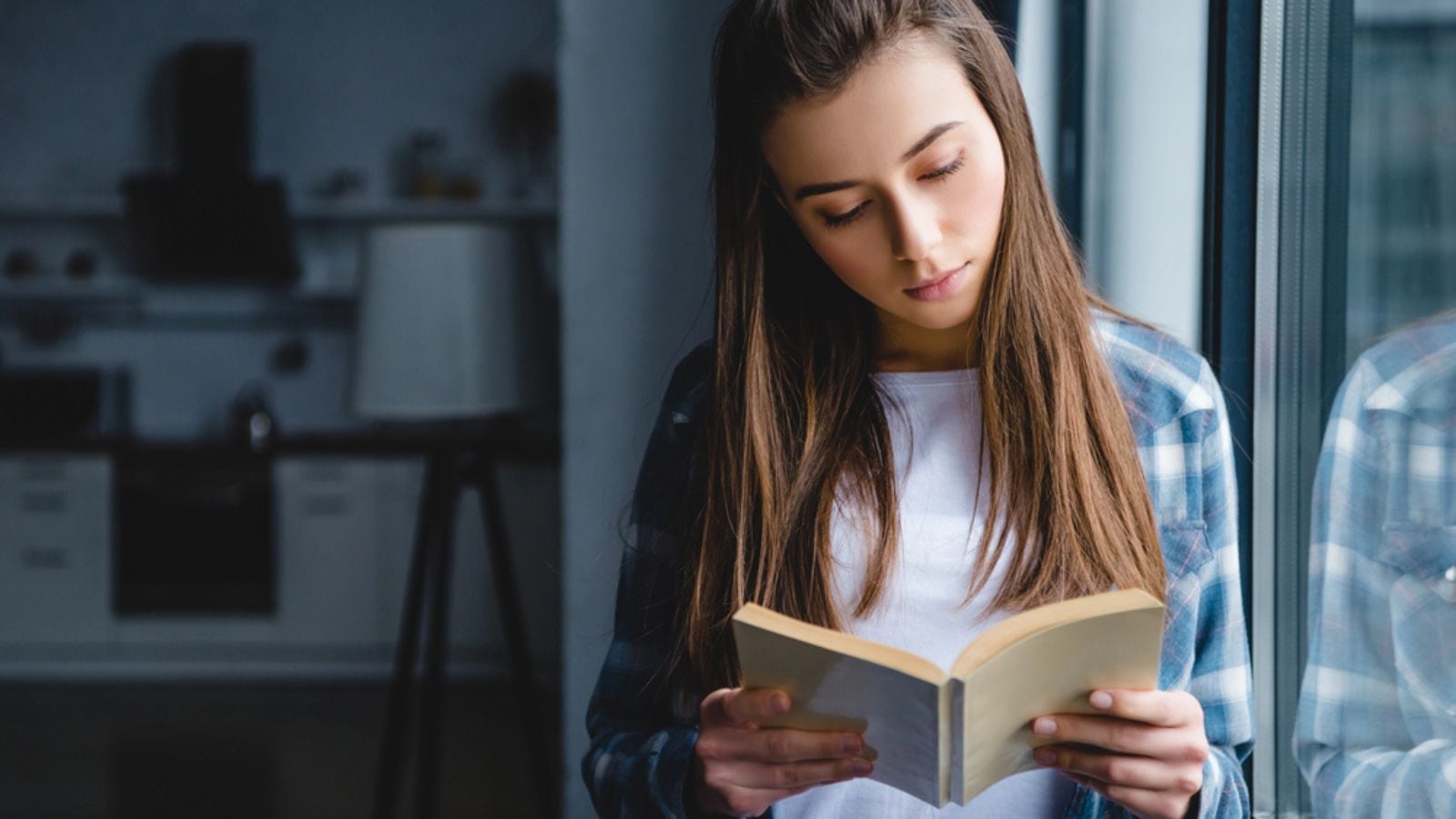 Beautiful young woman in checkered shirt standing near window and reading book