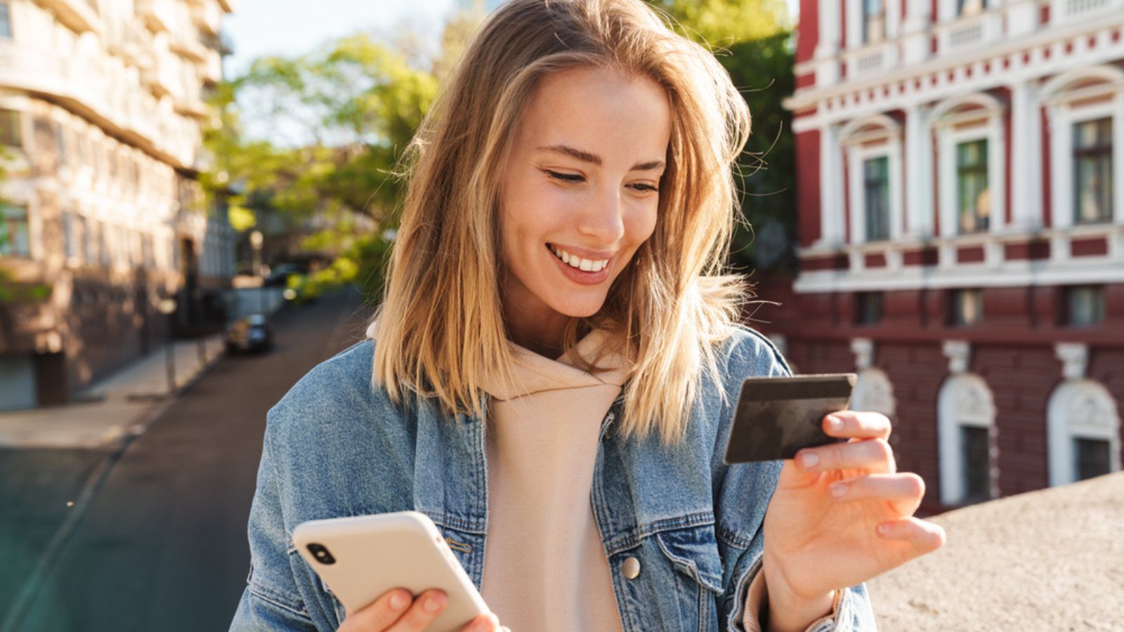 Cheerful young blonde girl shopping online with mobile phone and credit card while standing on a street