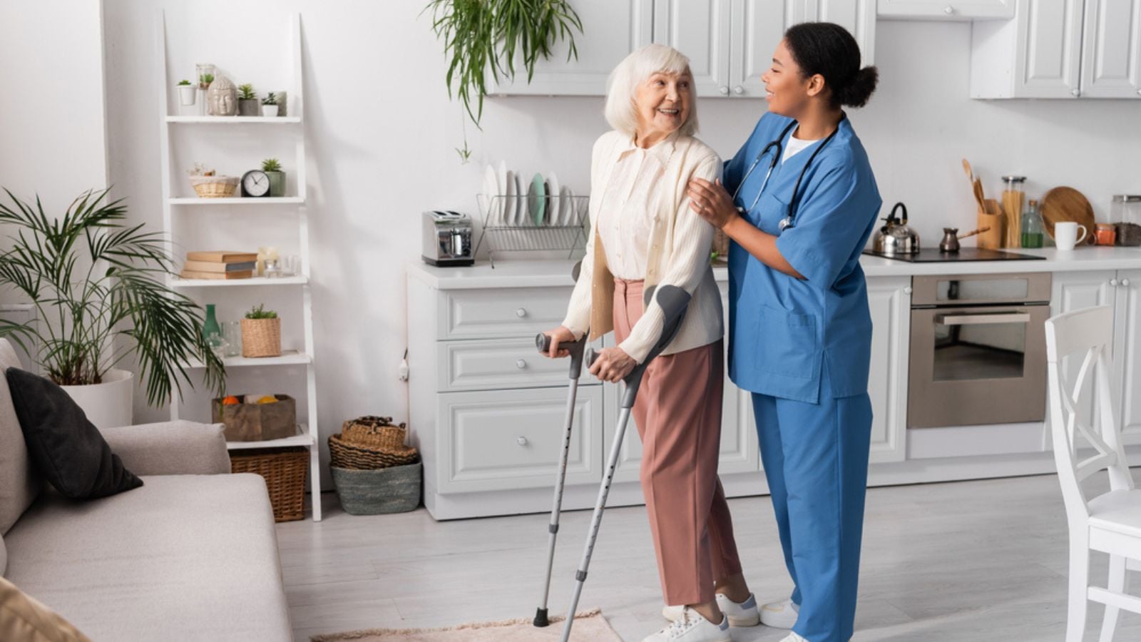 Full length of cheerful senior woman with grey hair using crutches while walking near multiracial nurse at home