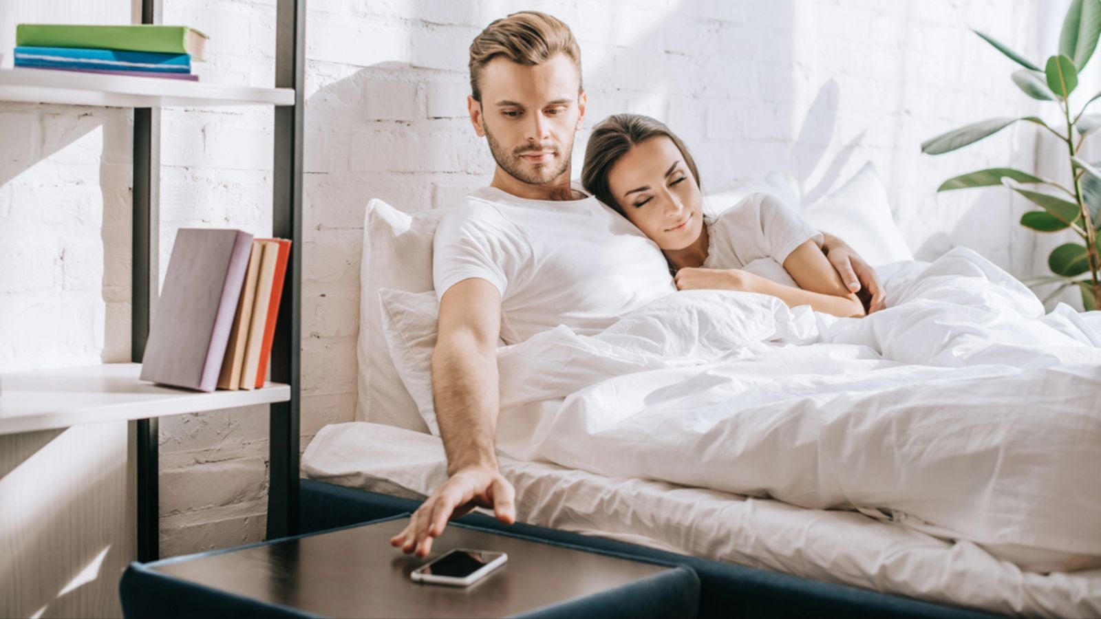 Handsome young man relaxing in bed with girlfriend and reaching for smartphone