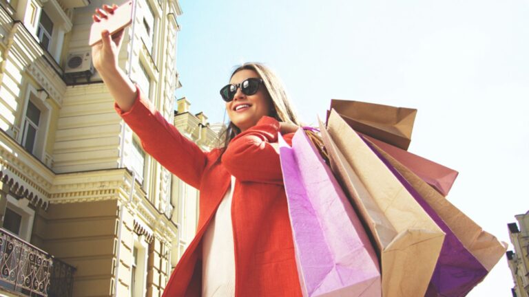 Selfie time. Portrait of cheerful and attractive woman in red coat and sunglasses taking a selfie with shopping bags