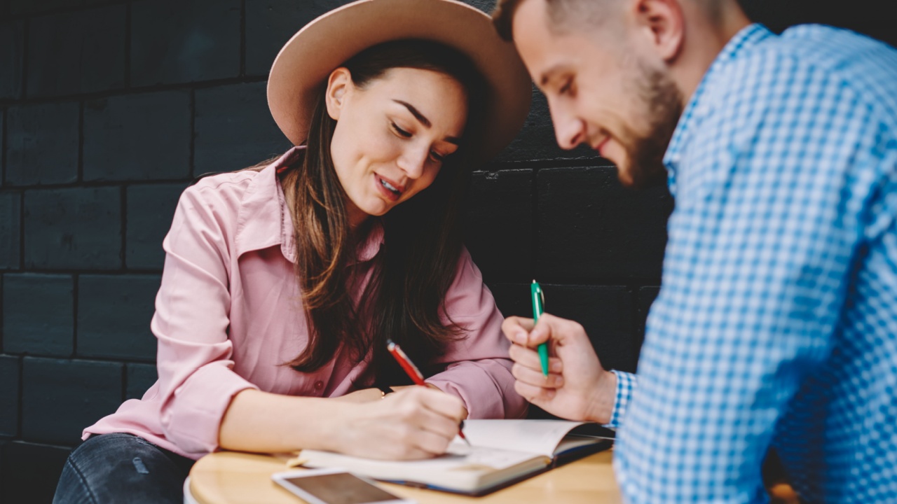 Couple in love dressed in casual wear discussing plan of common rest and writing down checklist in notepad spending leisure time in modern coffee shop.Young marriage noting list to do in notebook