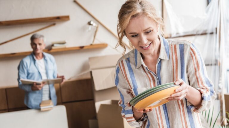 smiling woman packing plates while husband with books standing behind during relocation