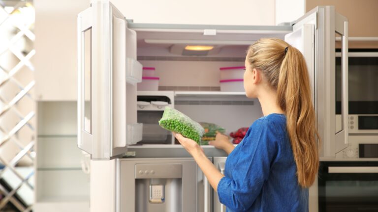 Young woman with frozen green peas near open refrigerator at home
