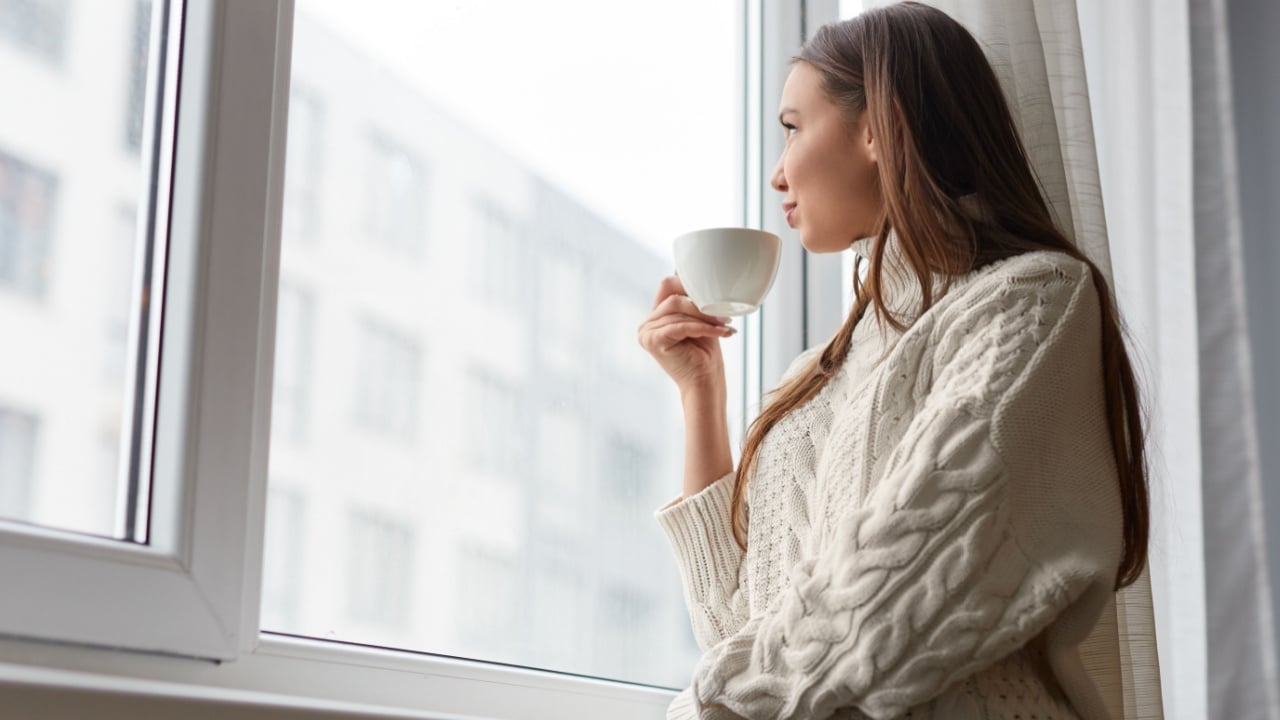 From below of wonderful dreaming woman in warm white sweater holding cup of aromatic hot drink looking away in window at home