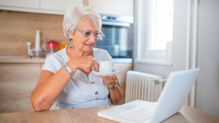 Elderly woman working on laptop computer, smiling, drinking tea. Senior woman using laptop. Elderly woman sitting at home, using laptop computer, smiling.