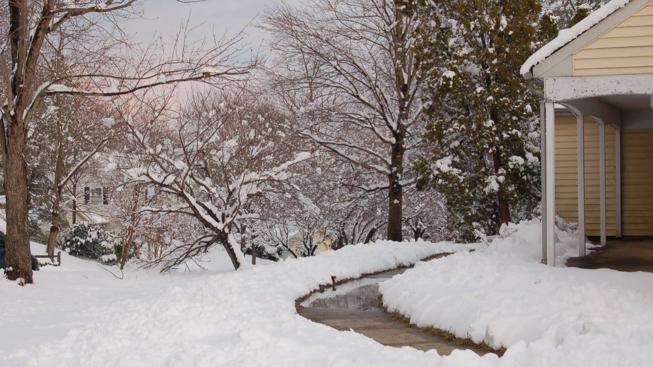Single family home with yard covered in snow during the winter. The sidewalk has been shoveled and iced over. The trees have a layer of snow on then. The sky is light blue and pink. No clouds.