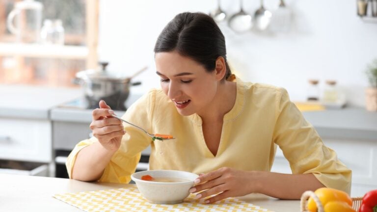 Young woman eating tasty vegetable soup at table in kitchen