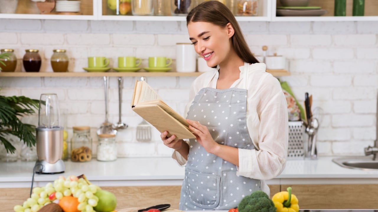 Girl reading cookbook in the kitchen, looking for recipe. Woman standing near table with fruits and vegetables