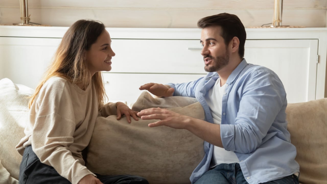 Smiling millennial couple sit on sofa in living room involved in interesting pleasant conversation at home, happy young man and woman talk chat relaxing on couch, enjoy romantic weekend or date