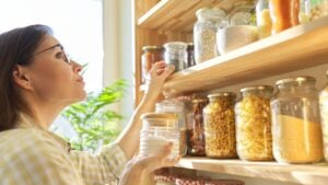 Food storage in pantry, woman holding jar of sugar in hand. Pantry interior, wooden shelf with food cans and kitchen utensils