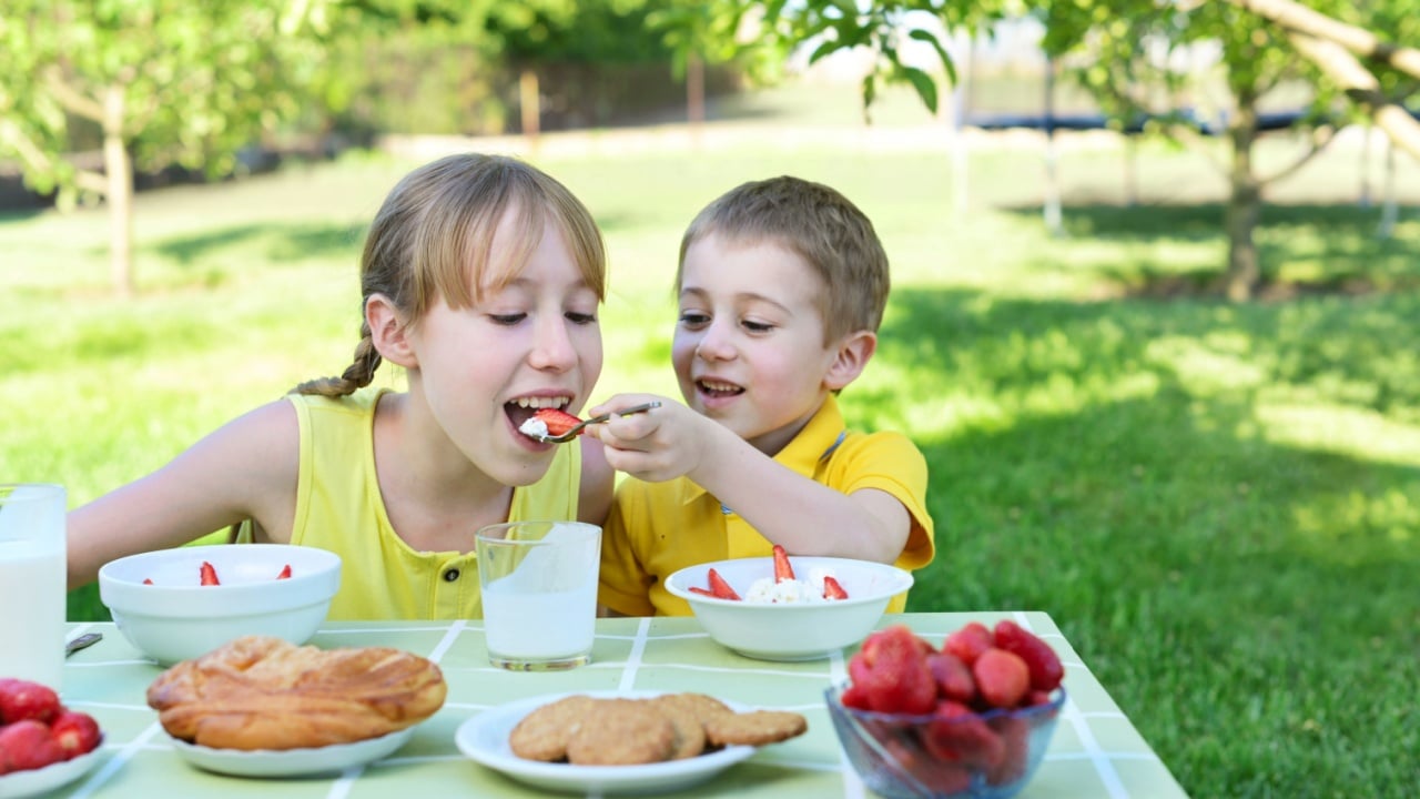 toddler brother treats sister healthy breakfast of dairy with fresh fruit strawberry, yogurt at a table. healthy eat concept