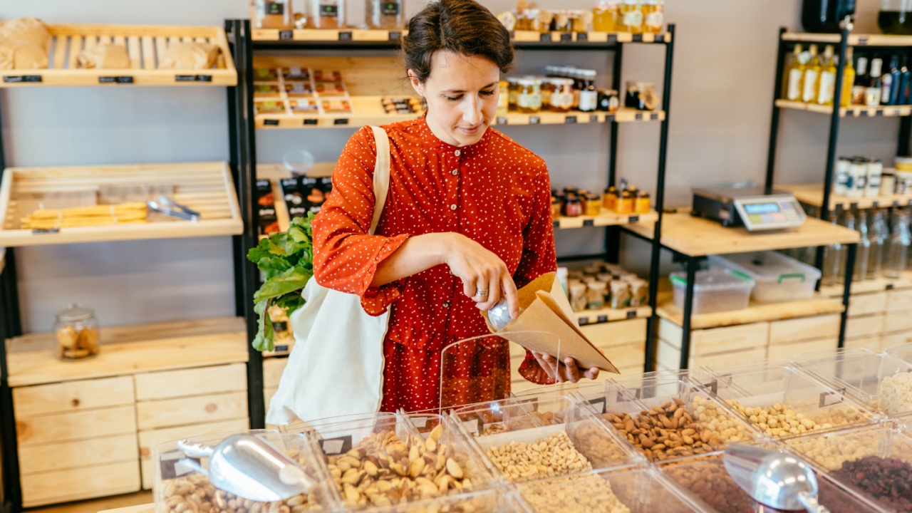 Young woman in red dress with reusable cotton bag doing shopping in plastic free store. Minimalist vegan girl buying nuts, groceries without plastic packaging in zero waste shop. Low waste lifestyle.
