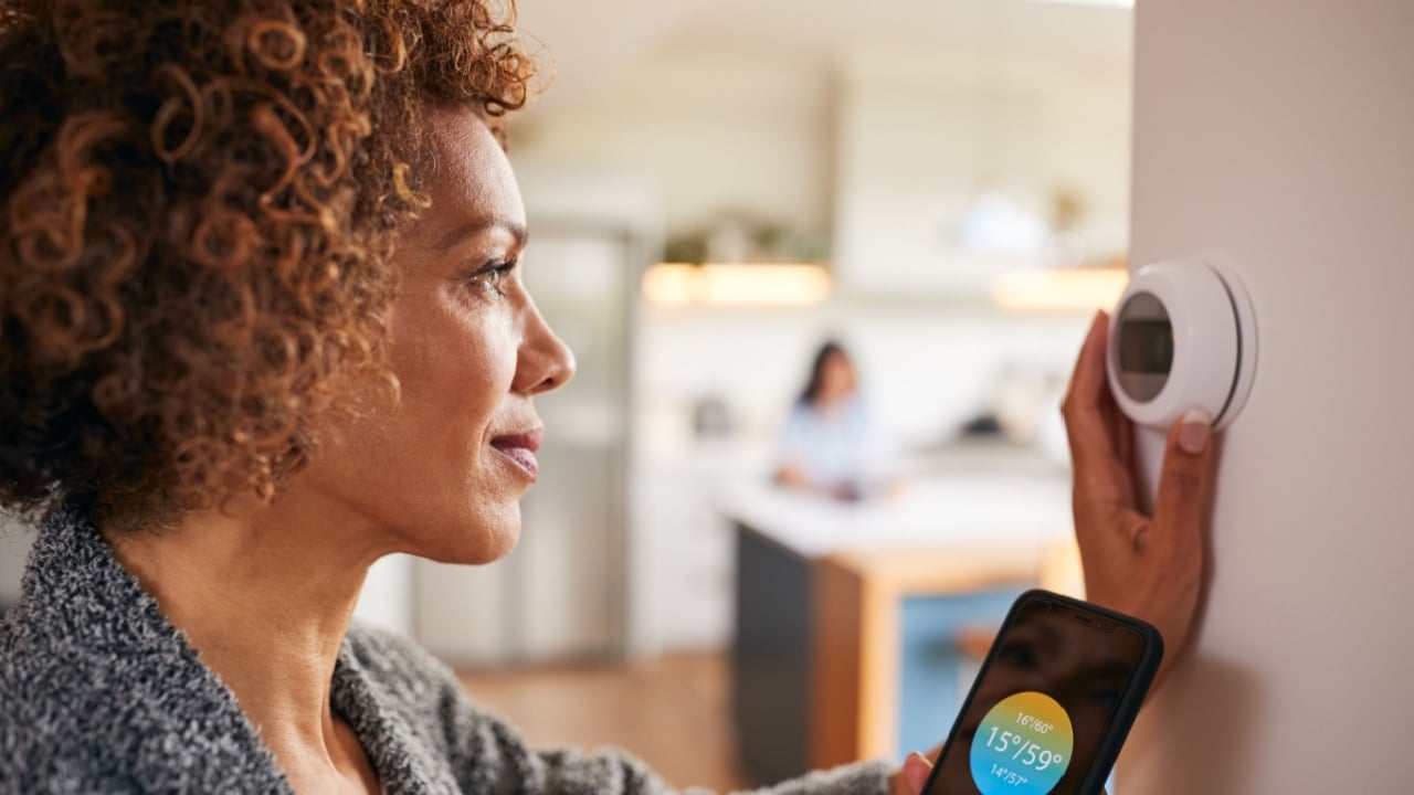 Mature Woman Using App On Phone To Control Digital Central Heating Thermostat At Home