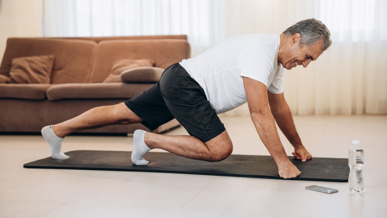 Elderly man doing mountain climber exercises on black yoga mat. Morning workout. Modern living room on background. Plastic bottle of water. Hard workout. Training at home. Elderly sport concept.