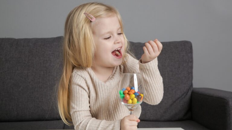 Portrait of adorable toddler girl eating colorful button-shaped chocolate candies at home.