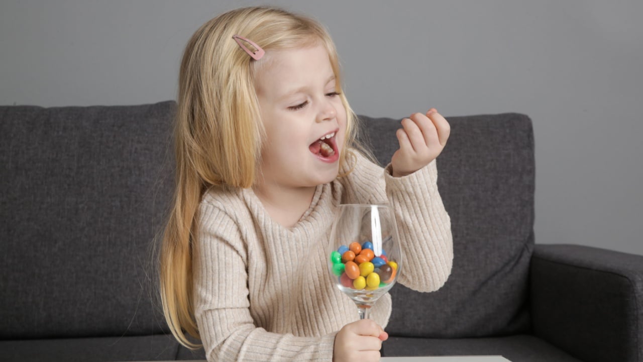 Portrait of adorable toddler girl eating colorful button-shaped chocolate candies at home.