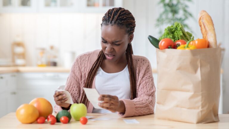 Expensive Groceries. Shocked African Housewife Sitting At Table In Kitchen, Checking Bills