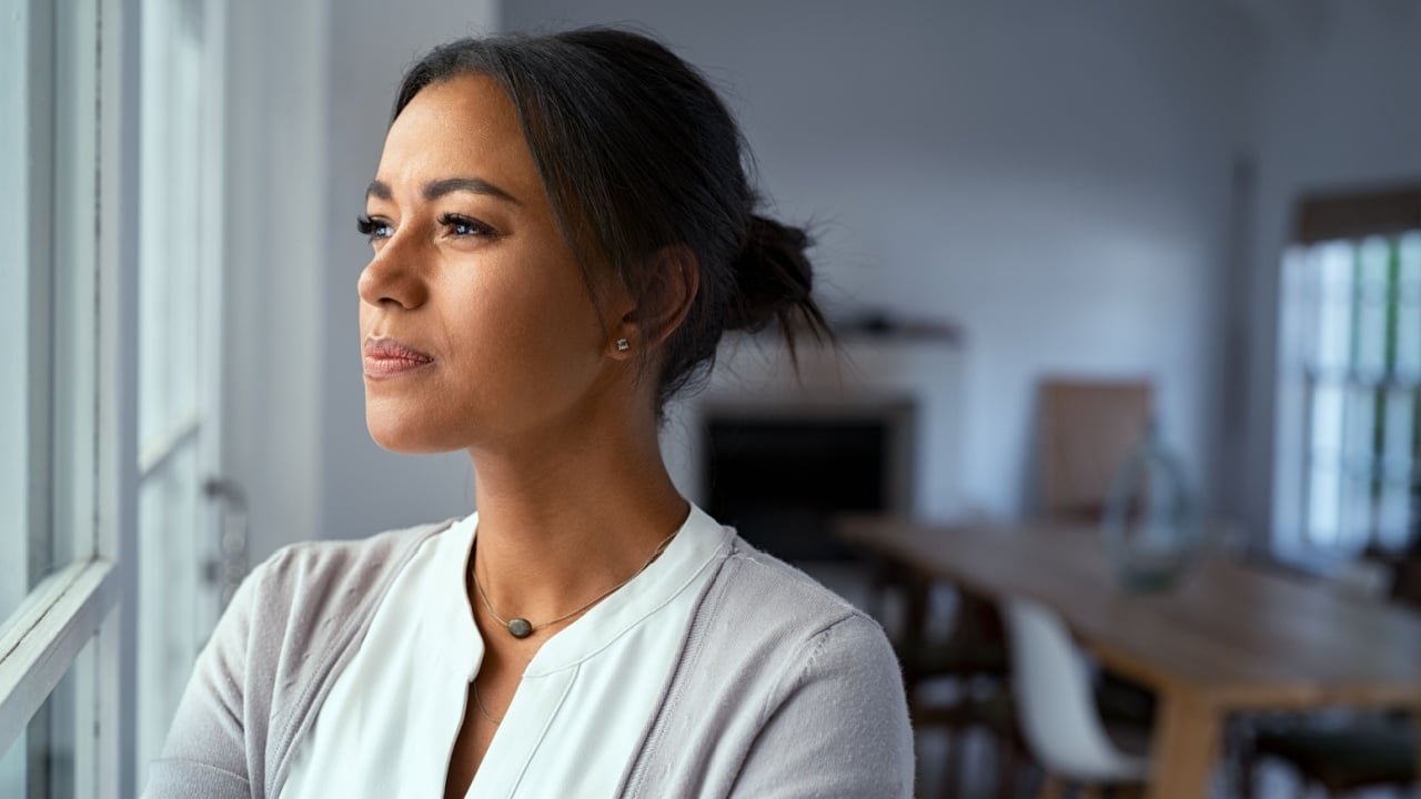 Mature african woman looking outside window with uncertainty. Thoughtful mid adult woman looking away through the window while thinking about her future business after pandemic. Doubtful lady at home.