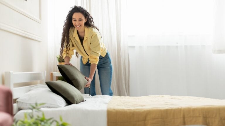 Cheerful young millennial lady making bed at home, holding pillow, doing housework in the morning. Positive stylish woman cleaning up her bedroom, everyday routine concept