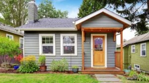 Small grey house with wooden deck. Front yard with flower bed and lawn