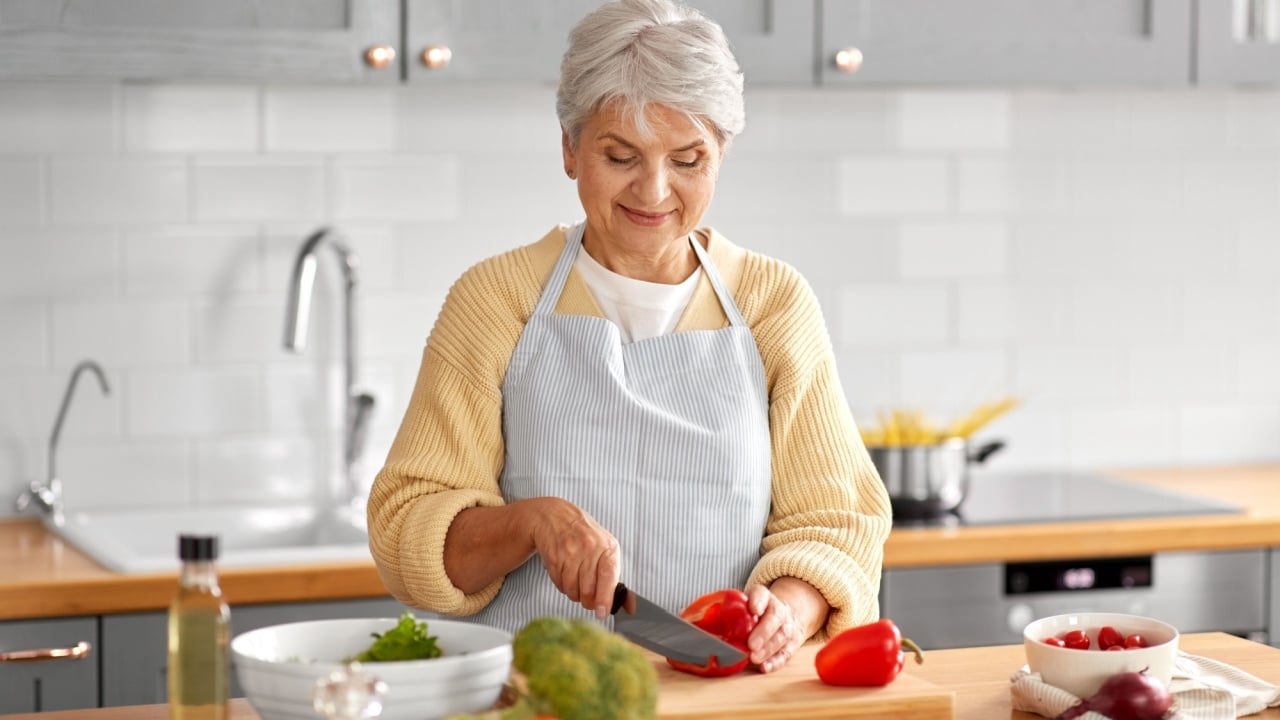 healthy eating, food cooking and culinary concept - happy smiling senior woman with knife chopping red pepper on kitchen at home