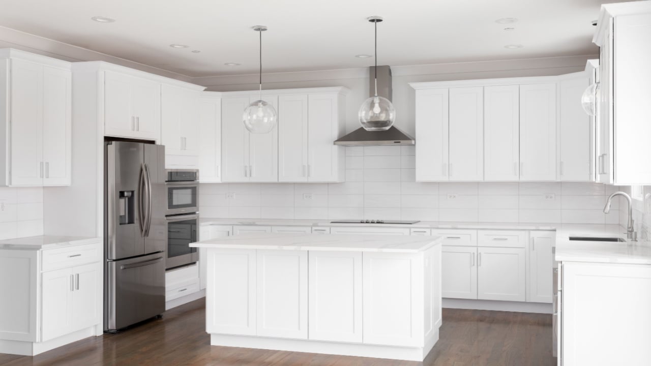 ELMHURST, IL, USA - JUNE 4, 2022: An all white, luxurious kitchen with pendant lights hanging over a large island, stainless steel appliances, tiled backsplash, and hardwood floors.
