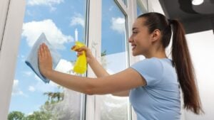 Happy young woman cleaning window glass with rag and spray indoors