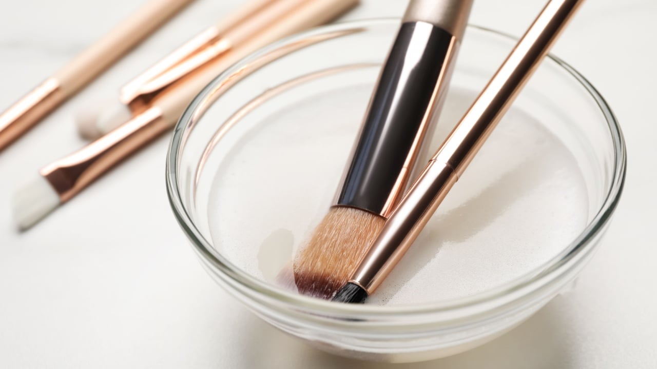 Clean makeup brushes in bowl with cleanser on white table, closeup