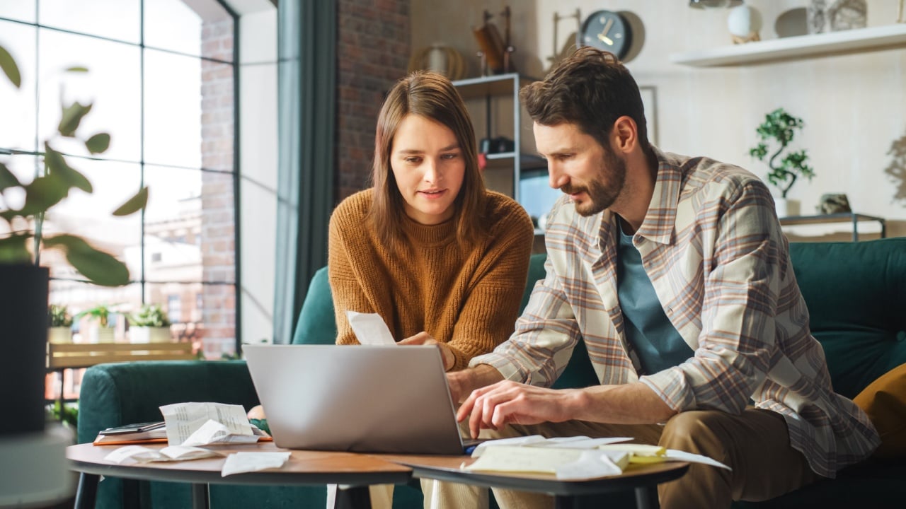 Stressful Accounting at Home: Couple Using Laptop Computer, Sitting on Sofa in Apartment. Young Family Filling Tax Forms, Mortgage Documents, Bills, Checks, Balances, Invoices are in Order