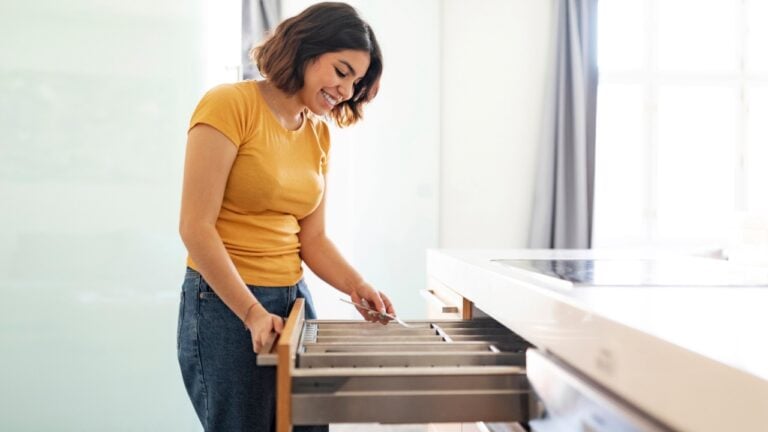 Young arab woman tidying up cutlery in drawer while cleaning in kitchen at home, happy middle eastern female assembling forks, enjoying making domestic chores, side view with copy space