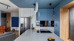Creative composition of kitchen interior with marble kitchen island, blue kitchen furnitures, gray fridge, modern lamp, bowl with fruits, big window and personal accessories. Home decor.