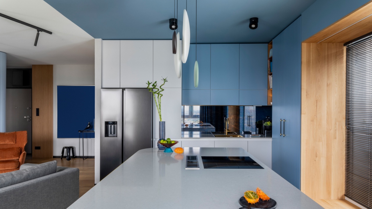 Creative composition of kitchen interior with marble kitchen island, blue kitchen furnitures, gray fridge, modern lamp, bowl with fruits, big window and personal accessories. Home decor.
