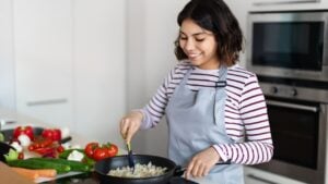 Beautiful young middle eastern woman holding cooking spatula, mixing rice with vegetables in frying pan on electric stove, preparing food at kitchen, copy space. Healthy diet, nutrition