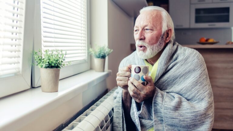 Elderly man in living room using heater at home in winter. Senior man warming his hands. Heating season. Cold adult man covered with blanket drinking tea at home.