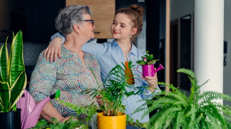 Two happy different generetion females enjoy, embracing, spending time together. Elderly grandmother teaches sharing experience cute teenage girl caring for a houseplant at home in the kitchen.