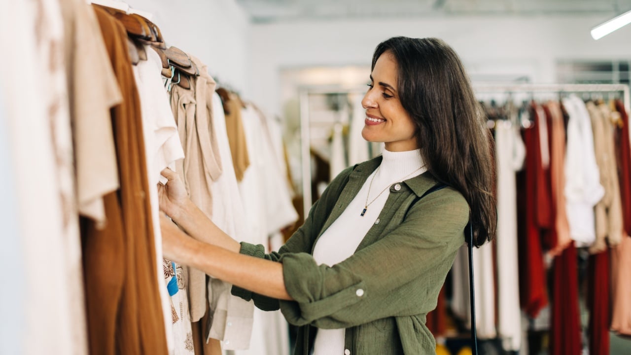 Happy woman stands in a fashion store, carefully choosing clothing items to buy. She browses through racks of stylish clothes, examining each one closely before making a decision.
