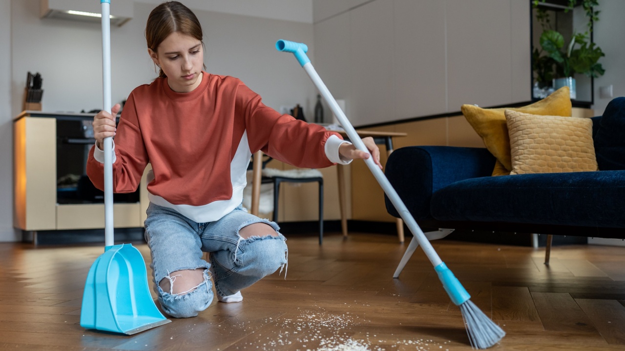 Independent teenager. Habit of cleaning house since childhood. Focused teen girl sweeps floor from scattered debris with broom with brush and dustpan. Adolescent do house chores, domestic cleanup.
