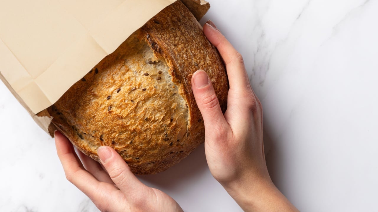 Fresh sourdough bread in woman's hands, top view. Woman takes a loaf of rustic wheat bread from a paper bag. Copy space banner on white marble background.