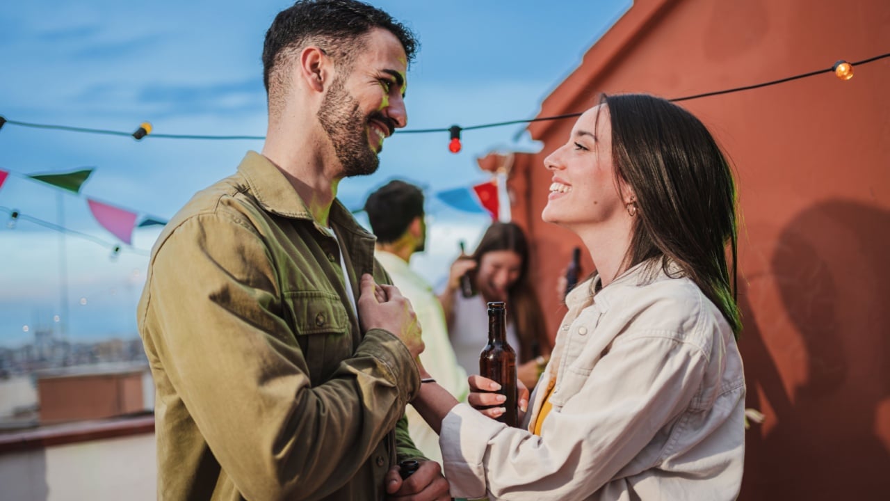 Smiling young couple flirting and having fun at a rooftop party with friends, drinking beer, enjoying and having a pleasant conversation. Handsome guy greeting his joyful girlfriend at celebration