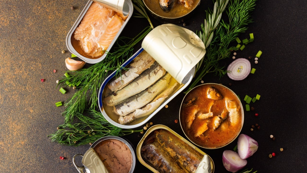 Different open tin cans with canned fish among spices and herbs on a brown background, canned salmon and mackerel, sprat and sardine, tuna and herring and fish pate, top view