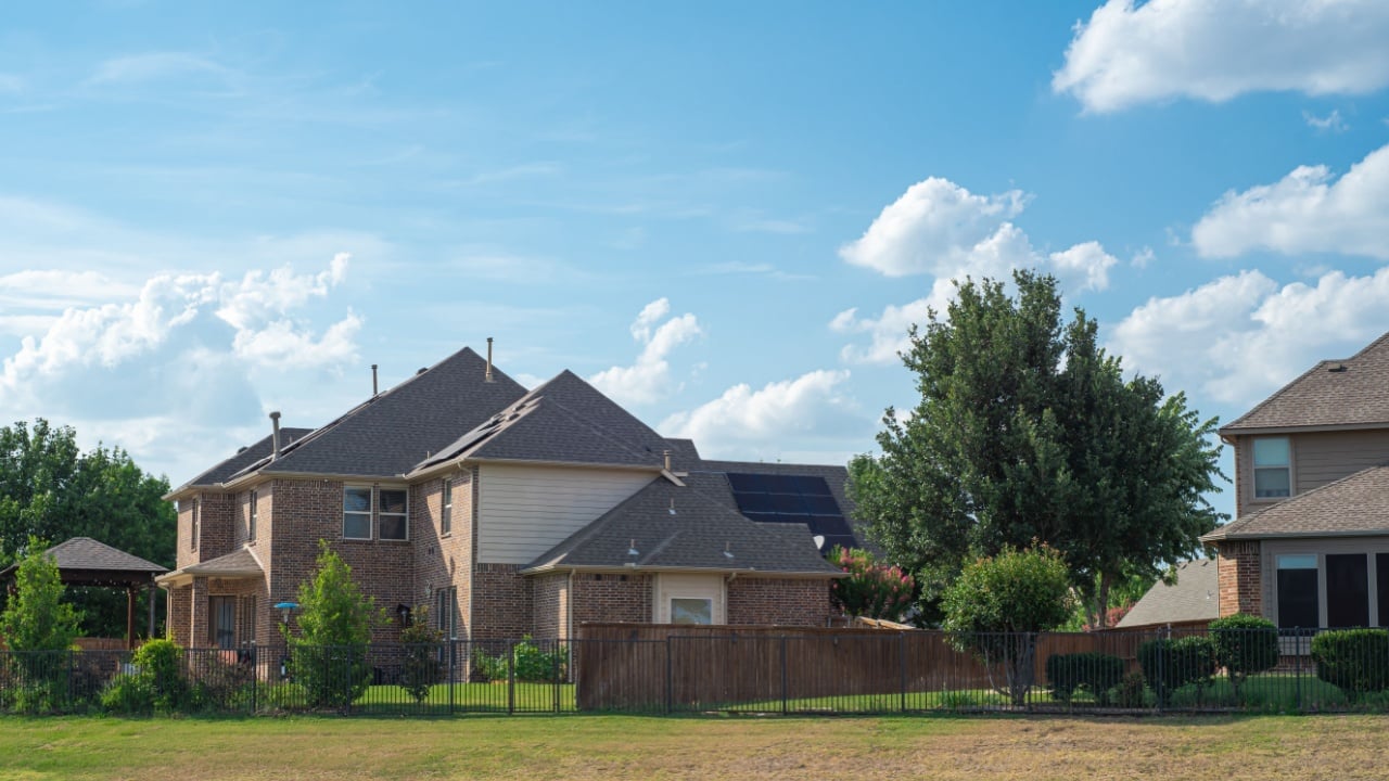 Backyard view of two-story suburban houses with solar roof panels, metal fence near large rolling hill grassy lawn of suburban park Flower Mound, Texas, USA. Upscale new development residential home