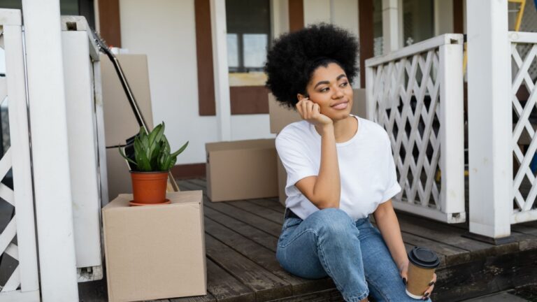 pleased african american woman holding coffee to go while sitting near carton boxes and new house
