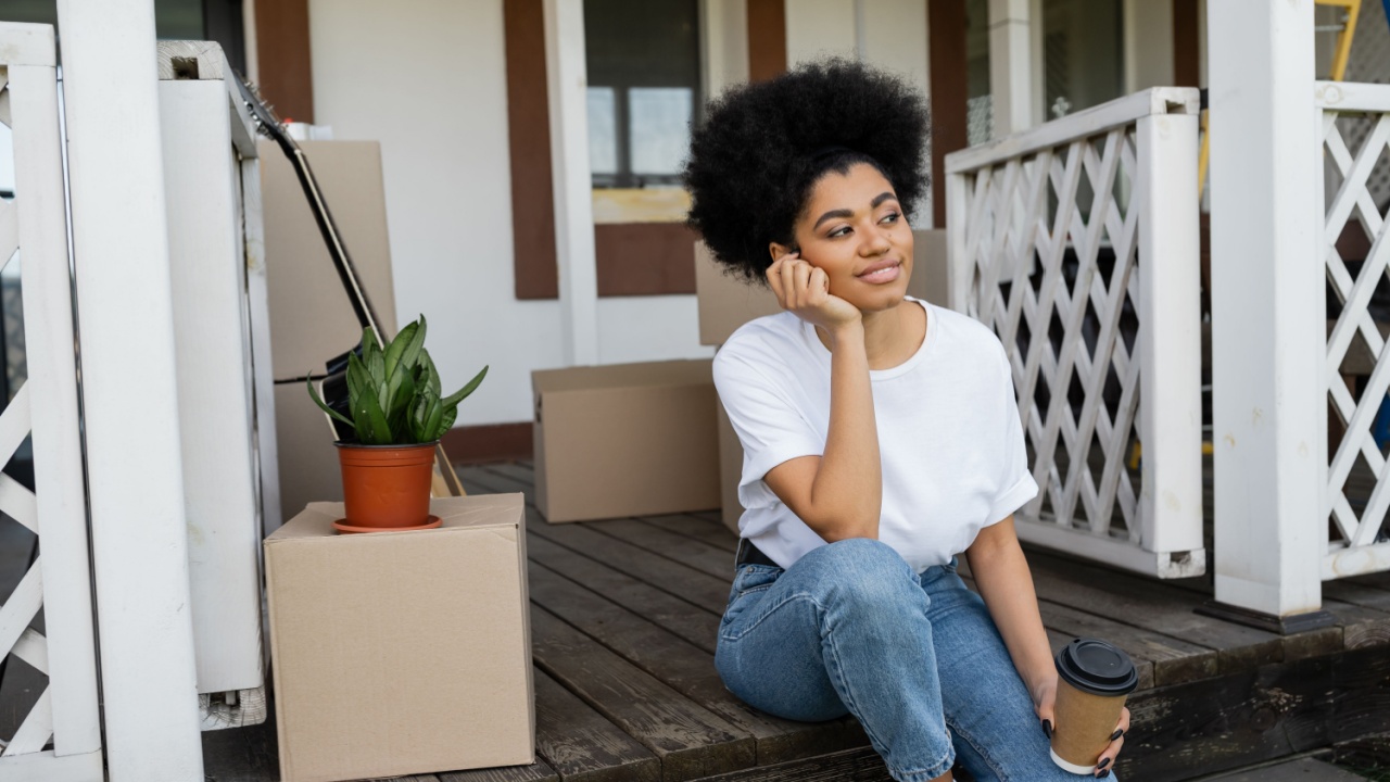 pleased african american woman holding coffee to go while sitting near carton boxes and new house