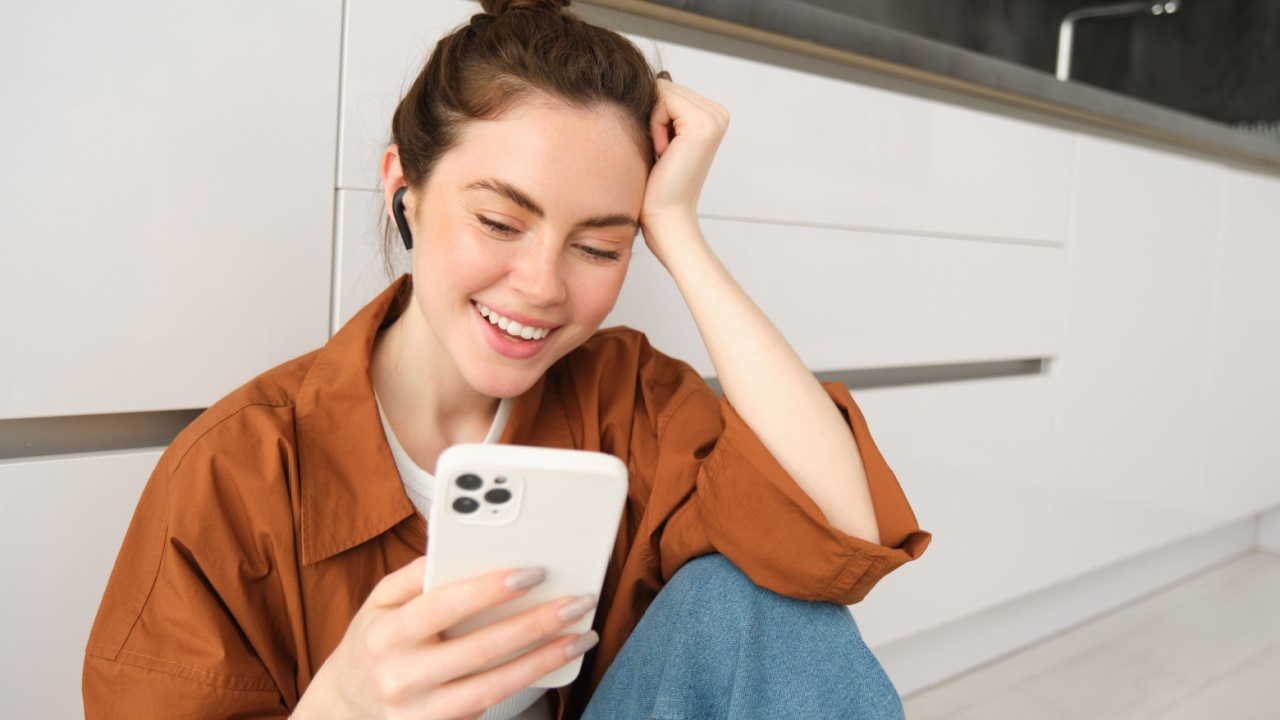 Portrait of young modern woman in wireless headphones, sitting on kitchen floor, using smartphone, making playlist with favourite songs, listens to music.