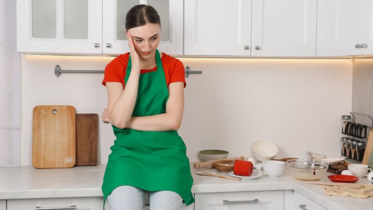 Upset woman sitting on countertop with dirty dishware and utensils. Mess in kitchen